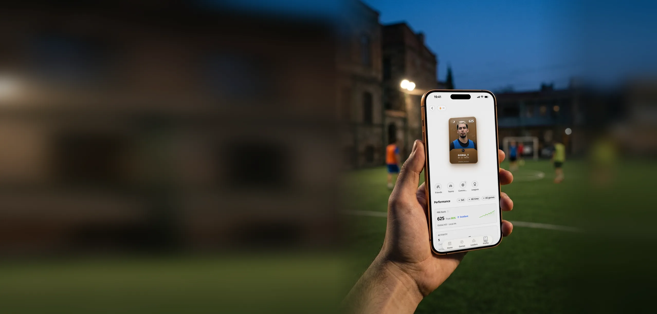 Player holding a phone with a Playbox player profile and PBX rank on a floodlit football pitch at dusk